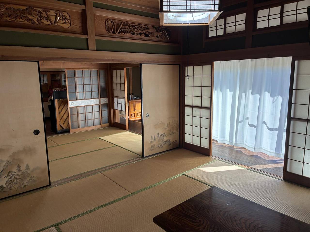 母屋の畳部屋。畳を敷いた続き間で、山水画の描かれた襖が開けてあり、その先にもう一間の畳の部屋が見える。天井近くの欄間には松や竹の彫り物、右手は障子とレースのカーテン越しに光の入る板張りの縁側