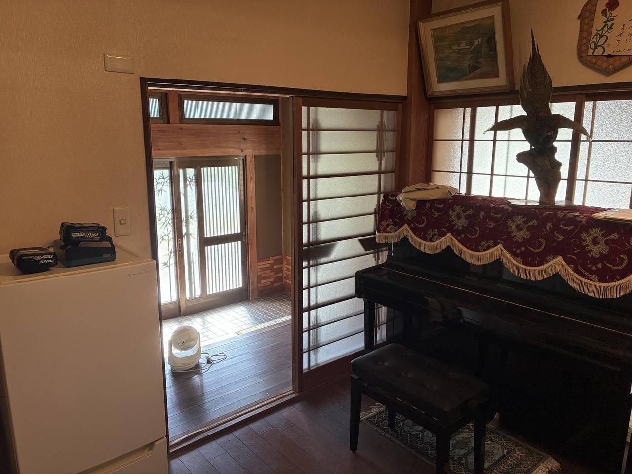 母屋の洋間。濃い色の板張りの床の部屋で、右手に覆いを掛けたピアノと椅子が置かれている。奥は障子と磨りガラスの窓、正面の引き戸の先に玄関の土間が見える