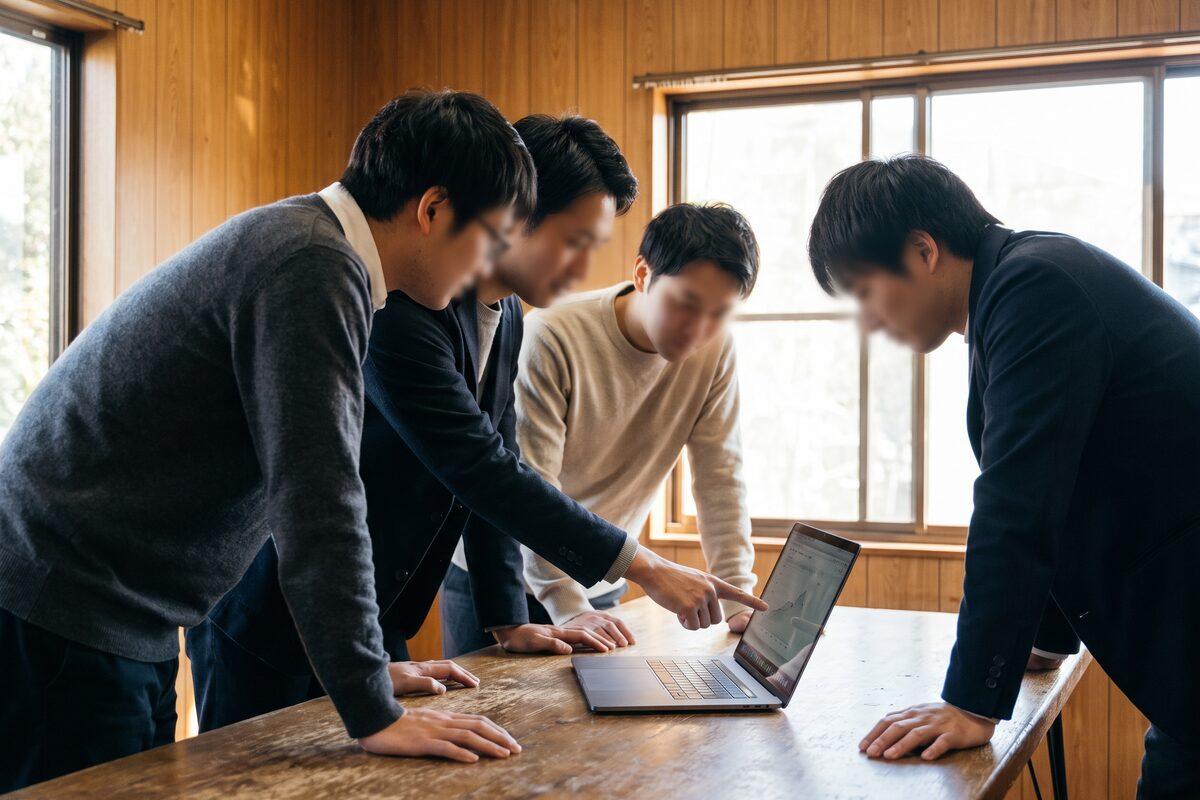 会議室の机で、1台のノートパソコンを4人の社員が囲み、1人が画面を指さしている社内勉強会の様子